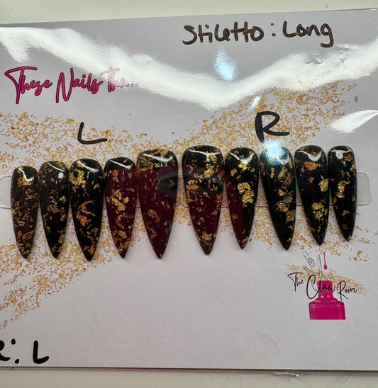 Long stiletto press-on nail set featuring a rich black and burgundy base with scattered gold foil accents. Nails are glossy and dramatic, exuding a luxe, edgy vibe.