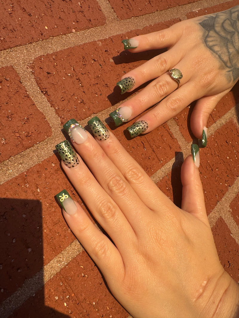 Close-up of a bold green and nude ombré press-on nail set with cheetah print accents, rhinestone details, and gold bow charms. Nails are displayed in stiletto and square shapes, giving off a wild yet glam vibe.
