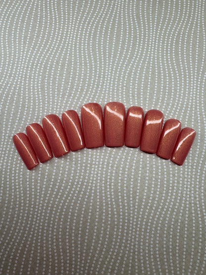 Set of medium square press-on nails in a vibrant coral shade with a golden shimmer finish, displayed on a branded card with glitter background.