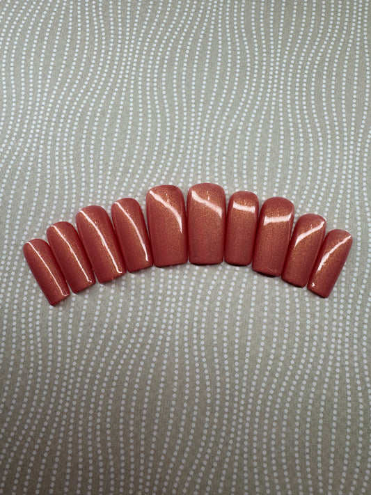 Set of medium square press-on nails in a vibrant coral shade with a golden shimmer finish, displayed on a branded card with glitter background.